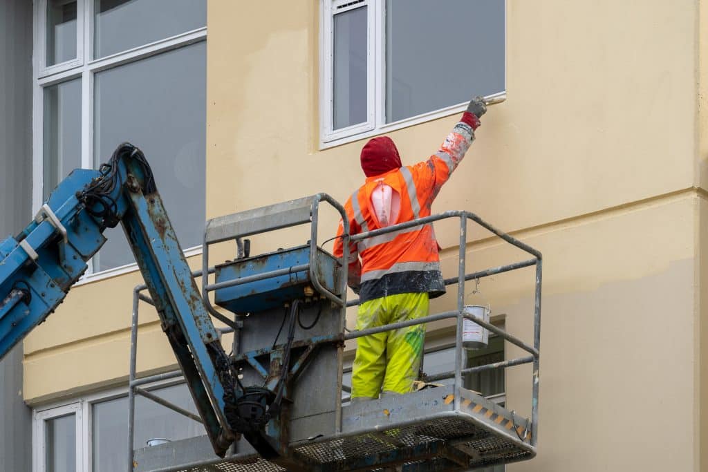 commercial exterior painting - A worker in hi-vis clothing hand paints the trim around a window in a pale sandy yellow color.