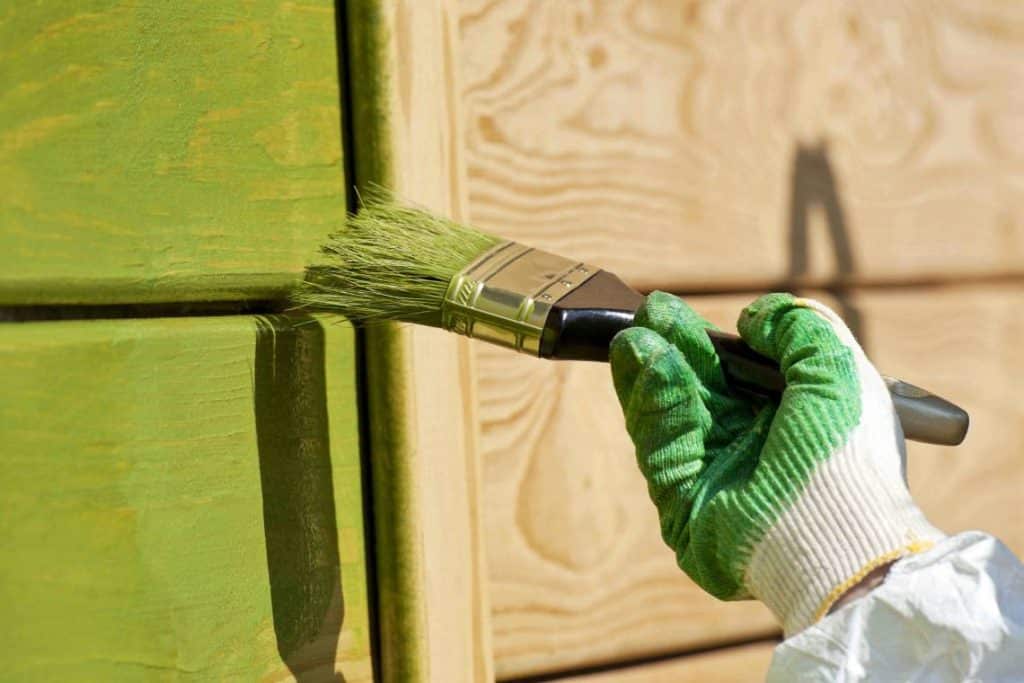 A painter's hand holding a paintbrush and applying green paint to wood siding.